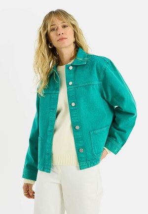 Femme aux cheveux blonds ondulés portant des boucles d'oreilles créoles dorées, une veste boutonnée bleu sarcelle sur un pull crème, et un pantalon blanc, debout devant un fond blanc.