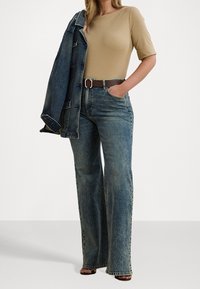 Top aderente beige chiaro, jeans flared in denim blu a vita alta, cintura con dettagli marroni e giacca di denim appoggiata sulla spalla.
