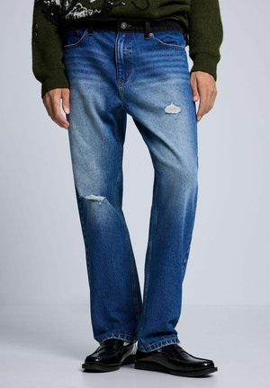 Uomo con jeans blu consumati, cintura nera e scarpe nere, in piedi davanti a uno sfondo semplice.