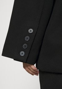 Mustassa ribbättynä bleiserissä on neljä tumman nappia, hienovarainen pinta ja istuva leikkaus, jossa käsi on asetettu sauman kohdalle.