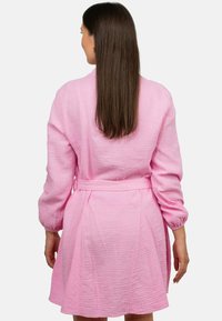 Abito rosa, con texture, a maniche lunghe e vita legata. Il tessuto ha una finitura stropicciata e arriva sopra il ginocchio.