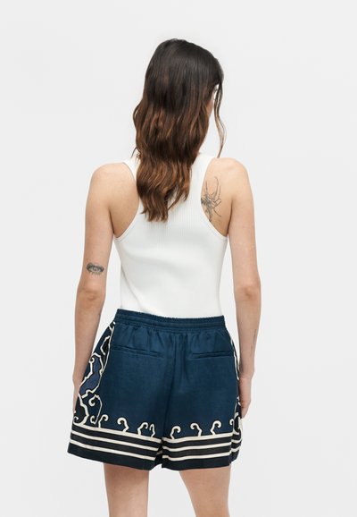 Γυναίκα με μακριά καστανά μαλλιά φοράει λευκό αμάνικο τοπ και σκούρο μπλε σορτς με λευκά διακοσμητικά σχέδια, εμφανίζεται από πίσω.