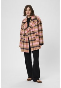 Abrigo cruzado de cuadros en rosa, marrón y negro, con grandes solapas con muescas y bolsillos delanteros. Llevado sobre una blusa negra y pantalones.