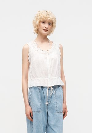 Γυναίκα με ξανθά σγουρά μαλλιά που φοράει λευκό αμάνικο μπλουζάκι με δαντέλα και βολάν, και ανοιχτό μπλε ψηλόμεσο τζιν παντελόνι με κορδόνι στη μέση.