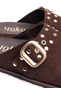 Sandales en daim marron foncé avec une grande boucle en or et des clous ronds en or sur la lanière, et semelle en caoutchouc noir.