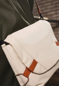 Beige canvas tas met een klep en sluiting, voorzien van bruine lederen accenten en zwarte hardware. Glad oppervlak met gestikte details.