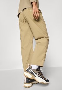 Beige lightweight trousers with a relaxed fit, paired with dark athletic shoes featuring a sturdy sole and orange accents.