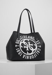 Czarna torba typu tote o fakturowanej powierzchni z dwiema długimi rączkami, z dużym białym okrągłym logo z napisem "GUESS LOS ANGELES CALIFORNIA 1981".