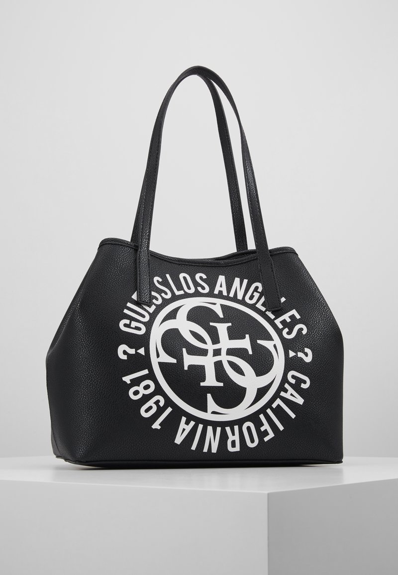 Czarna torba typu tote o fakturowanej powierzchni z dwiema długimi rączkami, z dużym białym okrągłym logo z napisem "GUESS LOS ANGELES CALIFORNIA 1981".