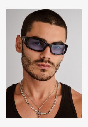 Occhiali da sole neri a forma rettangolare con lenti scure, dotati di un accento dorato sulla montatura. Indossati da una persona con una canotta nera, con collane d'argento visibili.