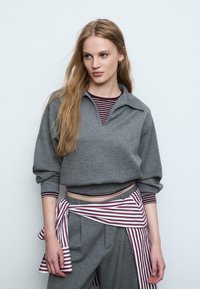Grå sweatshirt med v-hals og lange ærmer, parret med en bordeaux og hvid stribet skjorte. Stribet bælte om taljen. Blødt stof.