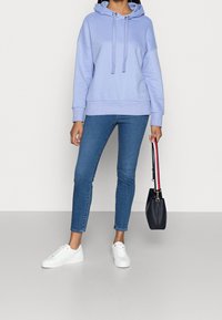 Felpa viola chiaro con cappuccio e stringa, abbinata a jeans skinny blu e sneakers bianche. Tieni in mano una piccola borsa nera con una tracolla colorata.