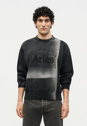 Νέος άνδρας με σγουρά μαλλιά που φοράει μαύρο και γκρι φούτερ με σχέδιο tie-dye, με το κείμενο «Aries» και αρχιτεκτονικό σχέδιο μπροστά.