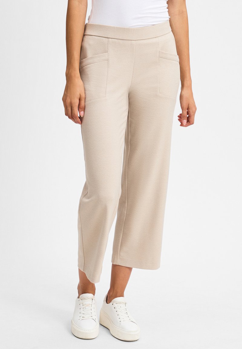 Beige weit geschnittene Hose aus weichem Stoff, mit zwei frontalen Taschen und einem glatten Bund, kombiniert mit weißen Sneakers.