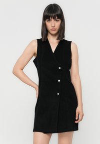 Robe sans manches en velours côtelé noir avec un col en V, présentant des boutons en argent sur le devant et une silhouette ajustée. Texture lisse sur l'ensemble.