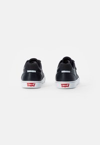 Ténis pretos de couro com solas de borracha brancas, detalhes cinzas e um logótipo vermelho da Levi's no calcanhar. A vista traseira revela um design elegante.