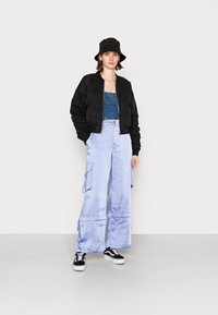 Casaco bomber preto, top cropped de ganga, calças cargo largas em lilás claro com bolsos, sapatilhas preto e branco, chapéu de balde preto.