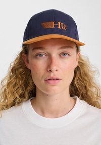 Navy blue cap with an orange visor. Features embroidered "HW" logo on the front, made of sturdy fabric. Classic structured shape.