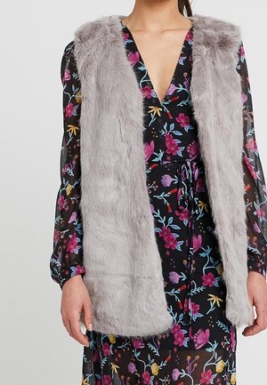 Vrouw draagt een mouwloos lichtgrijs nep bontvest over een zwarte bloemenjurk met lange mouwen en een diepe V-hals.