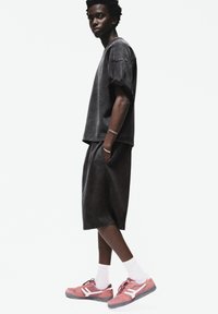 T-shirt oversize in denim nero, pantaloni shorts abbinati e sneaker rosa con accenti bianchi. Il modello si trova di fronte a uno sfondo bianco semplice.
