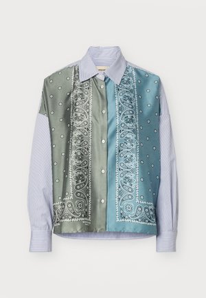 Jeune femme debout de face, portant une chemise boutonnée à motif vert et bleu, un pantalon bleu à jambes larges et des chaussures noires.