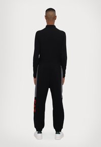 Tuta nera con colletto alto, caratterizzata da pannelli laterali grigi e un grande logo rosso sulla gamba. Il tessuto appare morbido e flessibile.