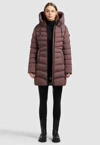 Mujer vistiendo un largo abrigo acolchado malva con capucha, leggins negros y botas negras, de pie contra un fondo claro.