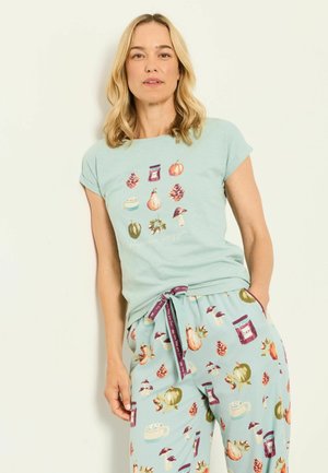 Frau trägt ein hellblaues Pyjama-Set mit herbstlichen Motiven wie Kürbissen, Kiefernzapfen, Pilzen, Gläsern und Tassen und posiert vor einem einfarbigen Hintergrund.