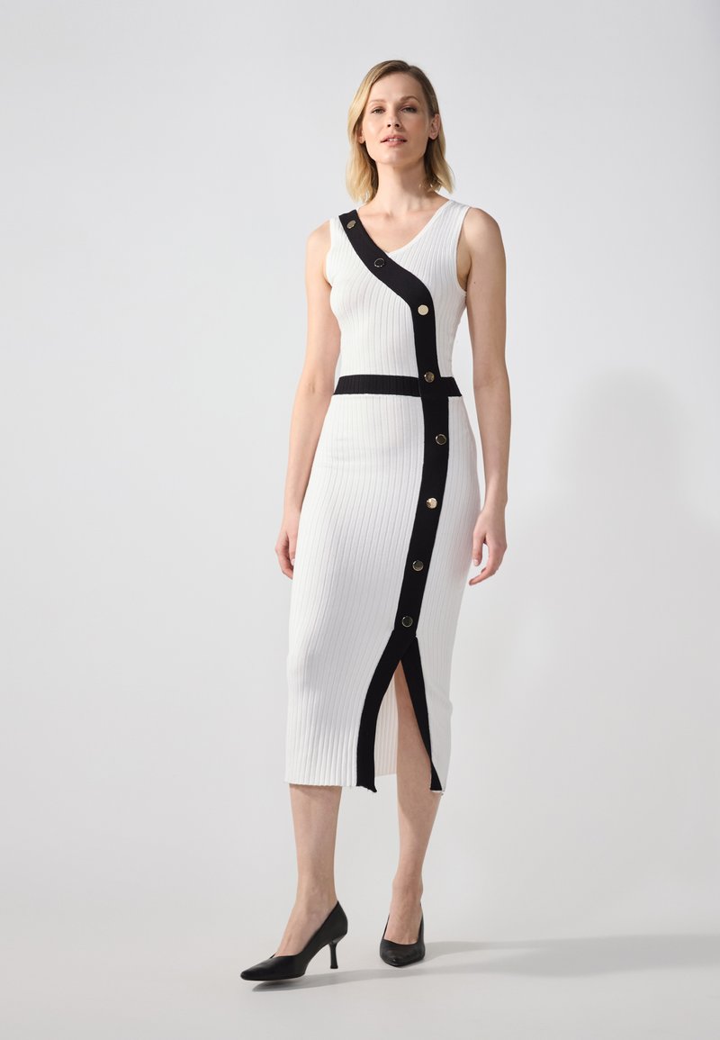 Biała, żeberkowana midi sukienka z czarnym asymetrycznym paskiem z złotymi guzikami, pionowym rozcięciem i dopasowaną sylwetką. Stylizowana z czarnymi szpilkami.