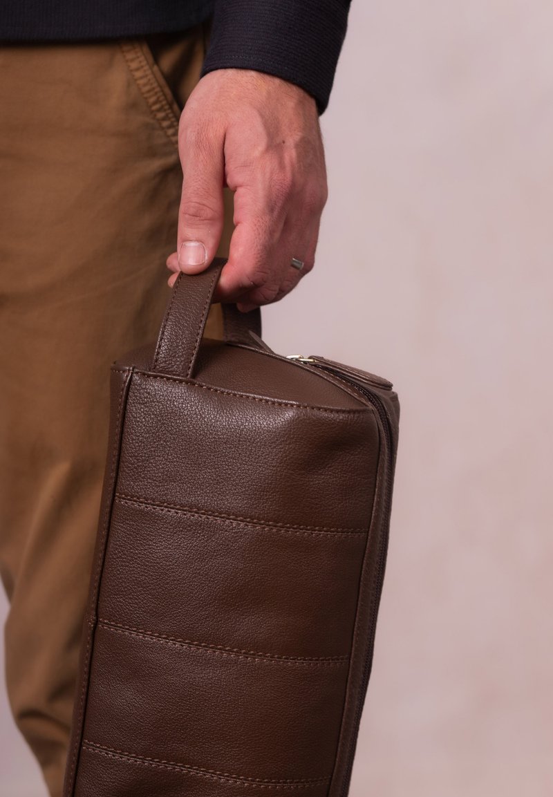 Hand holding a brown leather zippered toiletry bag against a beige background, wearing tan pants and a dark long-sleeve shirt.