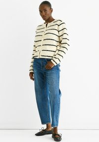 Striped cardigan in cream and black, textured fabric, round neckline, button closure, paired with blue straight-leg jeans and black flats.