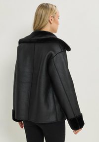 Giacca in ecopelle nera con un morbido colletto e polsini in pelliccia nera. Presenta una vestibilità comoda e dettagli di cucitura visibili sulla schiena.