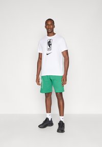 Fehér Nike póló az NBA logójával, zöld nadrággal, melyen oldalsó felirat található. Fekete sportcipő fehér zoknival egészíti ki a sportos megjelenést.