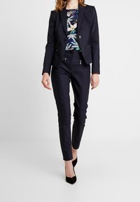 Mekko navy-värissä, jossa on viistolapelli, vetoketjulliset taskut ja kontrastivärinen kukkakuosisia paita. Yhdistetty tummat, hyvin istuvat housut ja mustat korkokengät.