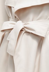 Beżowy płaszcz trenchowy wykonany z materiału o szerokim kołnierzu i związanym w talii pasku. Gładka tekstura, podwójnie szyte szwy i czyste linie w designie.