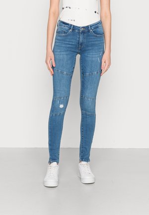 Slim fit blauwe jeans met gestikte horizontale paneeldetails en lichte slijtage aan de linkerknie, gedragen met witte sneakers en een wit shirt.