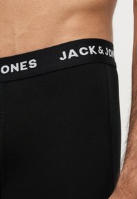 Taille masculine portant un sous-vêtement noir Jack & Jones avec le nom de la marque visible sur la ceinture, sur un fond neutre.