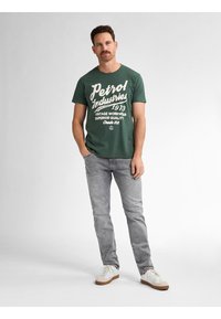 Zelené bavlněné tričko s bílým textem, kombinované se světle šedými džíny a bílými teniskami. Tričko má vintage grafický design.