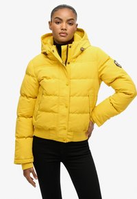 Gul pufferjakke med hætte, der har syede paneler, trykknapper og sidelommer. Materialet er glat med en mat finish.