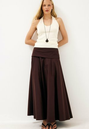 Blonde vrouw met een witte mouwloze top, lange donkerbruine rok, zwarte hangersketting en zwarte sandalen, staande tegen een witte muur.