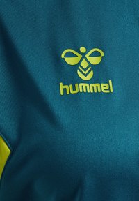 Turkoois stof met een groen logo met een bijenontwerp en het woord "hummel" eronder. Soepele textuur, minimale stiksels en contrasterende kleuraccenten.