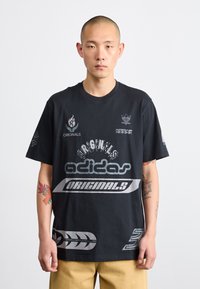 Melns kokvilnas T-krekls ar dažādiem Adidas logo, baltajiem un pelēkajiem grafiskajiem elementiem, kā arī īsām piedurknēm. Redzami ievērojami tetovējumi modeļa rokā.