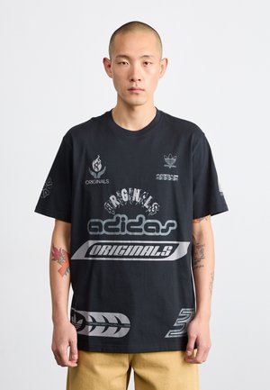 T-shirt in cotone nero con vari loghi Adidas, grafiche in bianco e grigio, e maniche corte. Tatuaggi notevoli visibili sul braccio del modello.