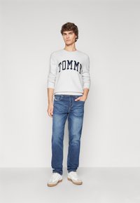Sivý pletený sveter s logom "TOMMY", modré džínsy a biele tenisky s béžovou podrážkou. Model stojí na jednofarebnom pozadí.
