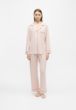 Jeune femme debout pieds nus portant un haut de pyjama rose clair à manches longues avec des boutons et un pantalon de pyjama large assorti.