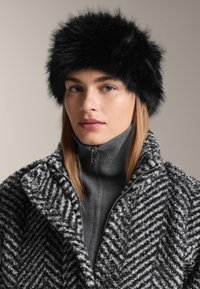 Čierna umelá kožušinová čiapka s objemným dizajnom. Nosená s textúrovanou sivou mikinou na zips s kosím vzorom.