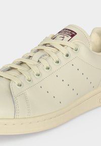 Αθλητικό παπούτσι Adidas Stan Smith εκρού με κρεμ κορδόνια, διάτρητες πλευρικές λεπτομέρειες και ετικέτα γλώσσας με λογότυπο σε σκούρο κόκκινο και λευκό.