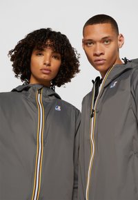 Vestes légères grises avec fermeture éclair à l'avant, dotées d'un détail de fermeture éclair rayé coloré et d'un logo en haut à gauche.