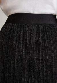 Černá plisovaná sukně s texturovaným, třpytivým povrchem a černým elastickým pasem s vyraženým názvem značky „JOOP!“ po obvodu vrchní části.