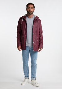 Mørkerød vandtæt jakke med hætte, trykknapper og to frontlommer, lagdelt over en grå sweatshirt, parret med lyseblå jeans og hvide sko.